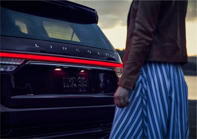 A person is shown near the rear of a 2023 Lincoln Aviator® SUV as the Lincoln Embrace illuminates the rear lights | Capital Lincoln of Wilmington in Wilmington NC